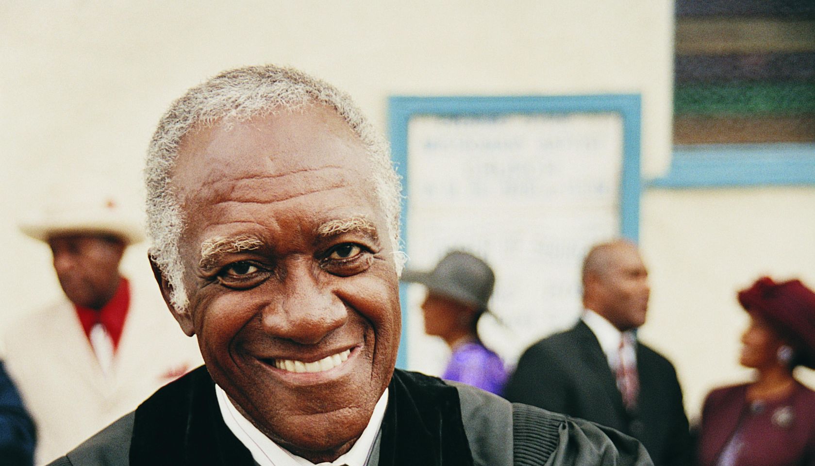 Portrait of a Smiling Priest Holding a Bible in His Crossed Arms With People in the Background