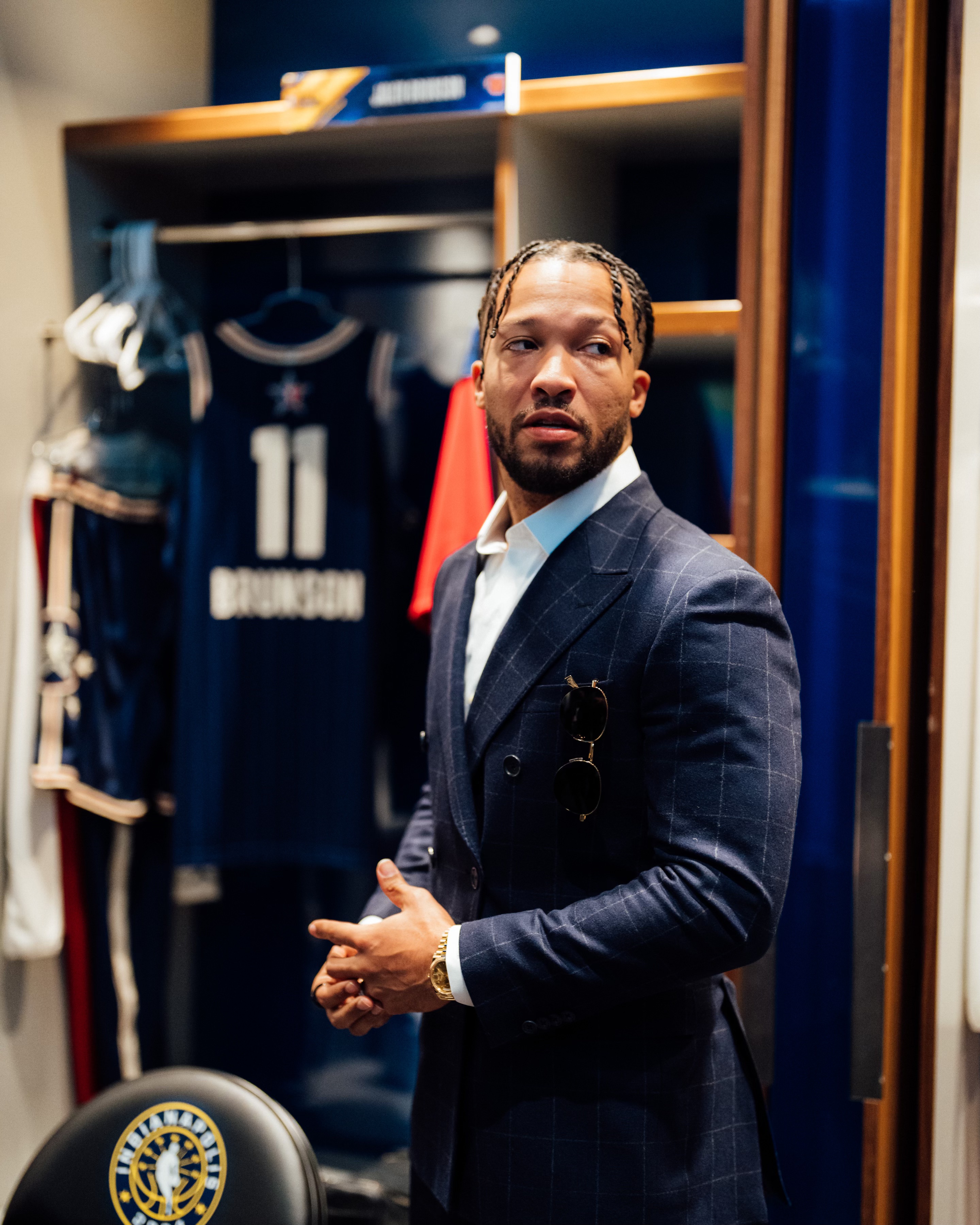 A well-dressed man with braided hair standing in a room with shelves and clothing racks, wearing a navy blue pinstriped suit and sunglasses.