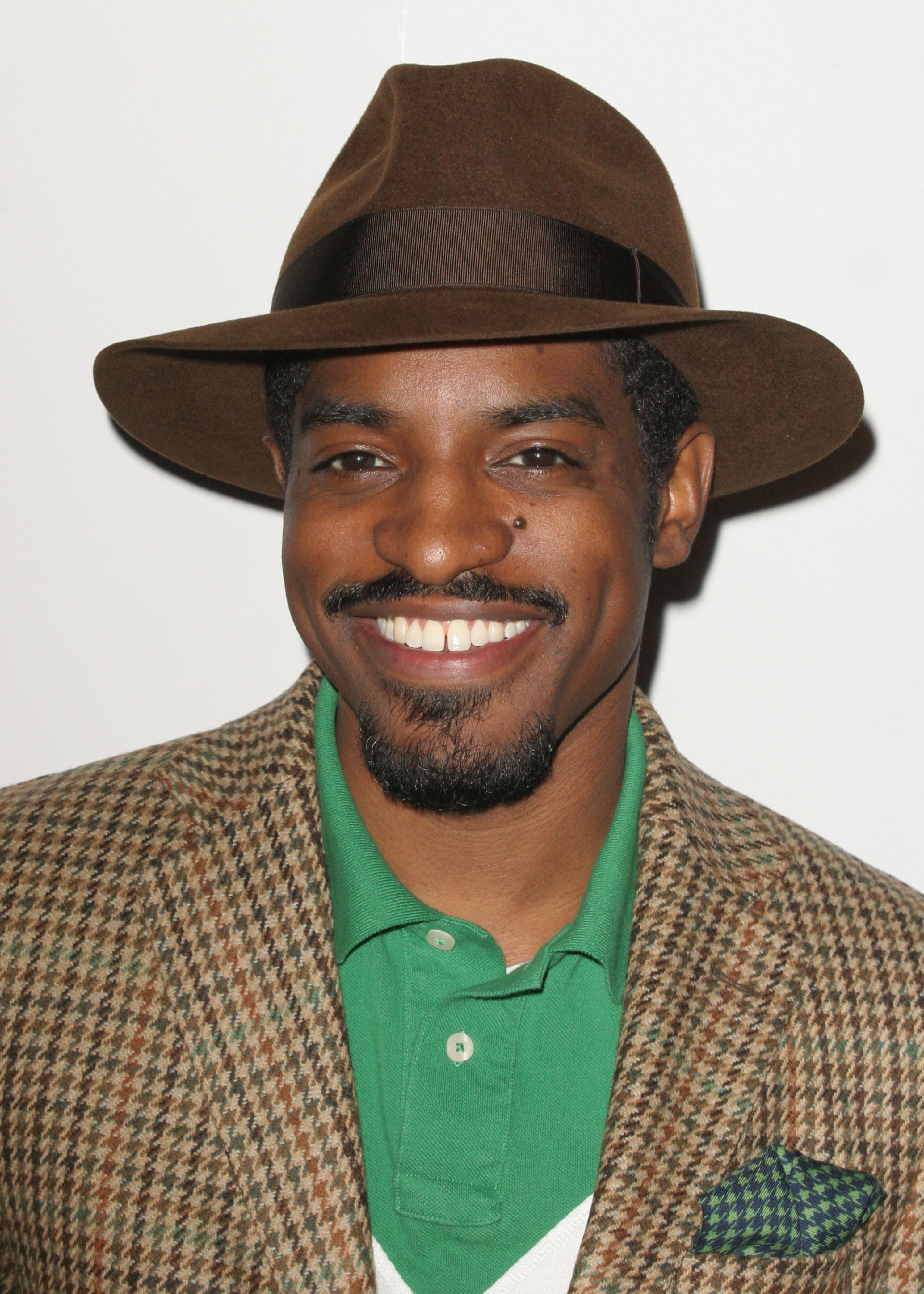 A smiling man wearing a brown hat and a tweed jacket over a green shirt.